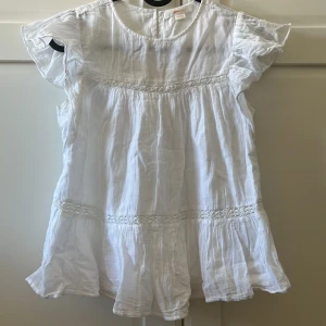 Vit blus med volang från Gina Tricot - Säljer en söt vit blus från Gina Tricot i storlek 146/152. Blusen har korta volangärmar och fina volangdetaljer längs nederkanten och midjan. Tillverkad i ett tunt och luftigt bomullstyg, perfekt för varma dagar. Använd få gånger.