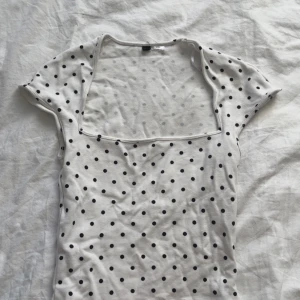 Vit prickig topp - Säljer en söt vit topp med svarta prickar. Toppen har en fyrkantig urringning och korta ärmar. Perfekt för en avslappnad stil.