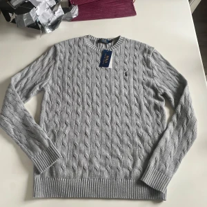 Ljusgrå kabelstickad tröja från Polo Ralph Lauren - Säljer en klassisk ljusgrå kabelstickad tröja från Polo Ralph Lauren. Tröjan har rund halsringning, långa ärmar och den ikoniska broderade loggan på bröstet. Perfekt för lager-på-lager och stilrena outfits. Helt ny storlek S men passar bättre till medium