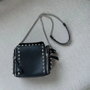 Svart axelväska med nitar och kedja - Cool svart axelväska från Zara i skinnimitation med silverfärgade nitar runt kanterna och detaljerade dragkedjor på sidorna. Väskan har en lång kedjerem som också är avtagbar🩶