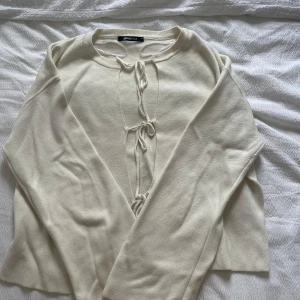 Vit stickad tröja med knyt från Gina Tricot - Säljer en vit stickad tröja från Gina Tricot med snygga knytningar framtill. Tröjan har lång ärm och en mjuk, bekväm känsla. Perfekt för lager-på-lager och enkel att styla till olika outfits.