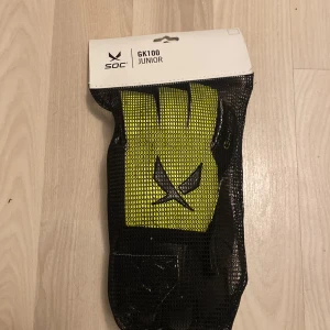 SOC GK100 Junior målvaktshandskar - Säljer ett par SOC GK100 Junior målvaktshandskar i svart och gul med justerbart kardborreband vid handleden. Handskarna har en cool hexagonstruktur på ovansidan och är perfekta för fotbollsmålvakter.