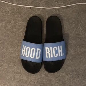 Blå och svarta slides från Hoodrich - Säljer ett par slides från Hoodrich med svart sula och blå rem med vit text. Perfekta för sommaren eller att ha hemma. Snygg och enkel design med tydlig logga på både ovansida och undersida. Använda 1,2 gånger