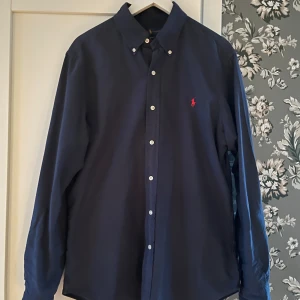Marinblå skjorta från Ralph Lauren - Säljer en klassisk mörkblå skjorta från Ralph Lauren med slim fit-passform. Skjortan har knappar hela vägen, button-down krage och den ikoniska röda loggan broderad på bröstet. Perfekt för dig som gillar stilrena plagg! Hör av er vid minsta fråga!