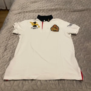 Vit piké från Polo Ralph Lauren med brodyr - Vit pikétröja från Polo Ralph Lauren med svart krage och detaljerad brodyr på bröstet. Tröjan har korta ärmar och knappar vid halsen. Tillverkad i 100% bomull och har klassisk polo-look med exklusiva emblem.