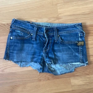 Blå jeansshorts med fransar från G-Star RAW - Snygga blå jeansshorts från G-Star RAW med råa, fransiga kanter och slitningar. Klassisk femficksmodell med knapp och dragkedja framtill. Perfekta för varma dagar och en avslappnad stil.