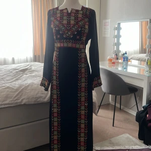 Svart palestinsk thobe långklänning ä - Elegant svart palestinsk långklänning med färgglatt broderi i rött, grönt och rosa. Klänningen har långa ärmar och ett traditionellt mönster som ger en unik touch. Perfekt för speciella tillfällen.