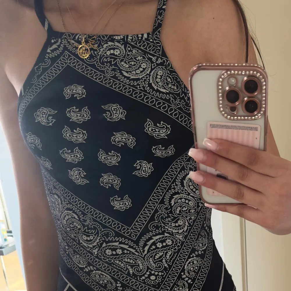 Säljer en svart klänning med vita paisley- och bandanamönster. Klänningen har smala axelband och är ärmlös med en kroppsnära passform. Perfekt för sommardagar eller festivaler. Storlek 38 men passar mig som har 32. Mekot.