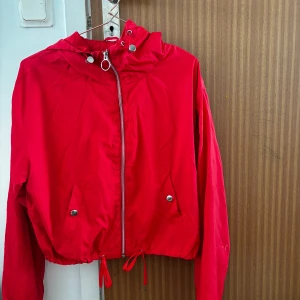 Röd vindjacka med huva - Säljer en röd vindjacka med huva och dragsko i midjan. Jackan har dragkedja framtill, två fickor med knappar och långa ärmar. Perfekt för blåsiga dagar och enkel att matcha med olika outfits.