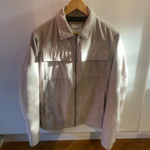 Beige mockajacka från Jack & Jones - Säljer en stilren beige mockajacka från Jack & Jones i storlek M. Jackan har dragkedja framtill, två stora bröstfickor med lock och klassisk krage. Jackan är i bra skick, inga synliga fläckar eller något sånt. Har däremot släppt lite i sömmen på höger bröstficka (se bild 4). 