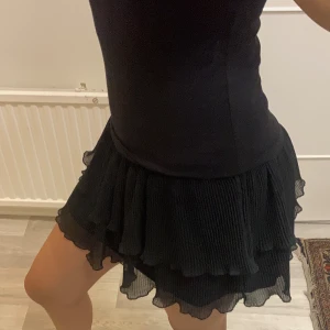Svart volangkjol ifrån Lindex - Snygg svart kortkjol med volanger som ger en lekfull look. Perfekt för att styla med olika toppar och accessoarer. Kjolen har en bekväm passform och är ett måste i garderoben för alla modeälskare.
