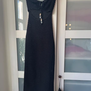 Svart strand klänning  - Svart virkad långklänning med snygg cut-out framtill och knytning vid bysten. Klänningen har dekorativa pärlor och snäckor som hänger ner från knytbandet. Perfekt för sommardagar eller strandhäng.