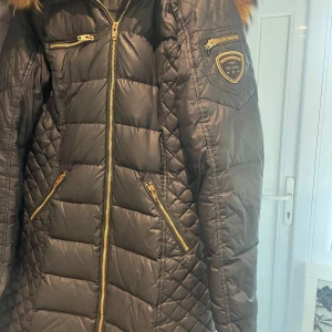 Svart dunjacka med fuskpäls från Rockandblue - Snygg svart dunjacka från Rockandblue med guldiga dragkedjor och stor huva med fuskpäls. Jackan har quiltad design, flera fickor och Rockandblue-logga på ärmen. Perfekt för kyliga dagar.