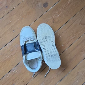 Valentino Garavani sneakers med blå detalj - Säljer ett par vita Valentino Garavani sneakers med bred mörkblå panel på sidan och blå snörhål. Skorna har rund tå och klassisk snörning. Perfekta för dig som gillar stilrena och exklusiva sneakers.