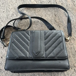 Svart axelväska från Steve Madden - Snygg svart axelväska från Steve Madden med quiltad design och justerbar axelrem. Väskan har guldfärgade metalldetaljer och magnetlås. Insidan är fodrad med logotypmönster och har flera fack för enkel organisering.