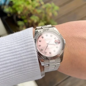Lyxig klocka med rosa urtavla - Elegant klocka med en glittrig rosa urtavla och silverfärgad boett. Armbandet är i matchande silvermetall och har en klassisk design. Perfekt för den som vill ha en stilren accessoar med en touch av färg.