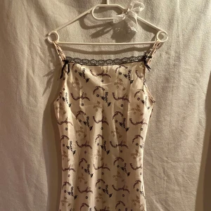 Beige satinklänning med spets och blommigt mönster - Säljer en söt beige satinklänning med tunna justerbara axelband och svart spetsdetalj vid urringningen. Klänningen har ett diskret blommigt mönster i brunt och svart samt små rosetter vid axelbanden.