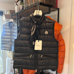 Moncler Gui Väst - Helt ny svart Moncler Gui med kvitto, tags, extra knappar och dustbag. Storlek 2 passar S/M, 2021 modell. Nypris 9200 mitt pris 4000kr! Över 250 deals gjorda med massvis av refs att visa, bara att skriva vid frågor eller funderingar!
