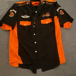 Svart och orange Harley Davidson skjorta - Cool svart och orange skjorta från Harley Davidson med broderade märken fram och bak. Skjortan har korta ärmar, två bröstfickor med lock och tydliga kontrastpaneler i orange. Lätt att styla och går att göra mycket med den! 