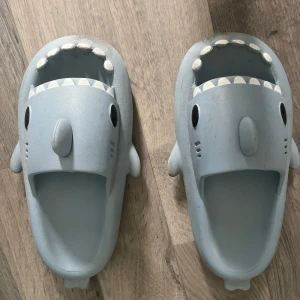Grå hajformade sandaler - Säljer ett par grå sandaler med hajdesign. Skorna har öppna tår och detaljer som tänder, fenor och ögon vilket ger dem en lekfull look. Perfekta för dig som vill sticka ut med roliga skor!