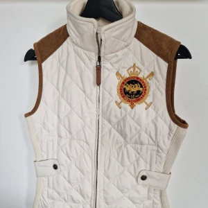 Vit quiltad väst från Ralph Lauren Sport - Snygg vit quiltad väst från Ralph Lauren Sport med bruna detaljer i mocka och broderat emblem på bröstet. Västen har hög krage, dragkedja framtill och markerad midja med knappar. Perfekt för en sportig och klassisk look. I väldigt bra skick