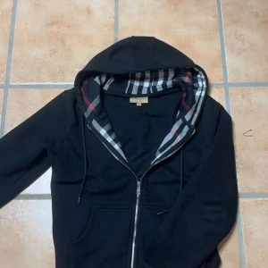 Burberry zip hoodie - Obs: liten i storlek. Snarare S. Säljer en stilren svart huvtröja från Burberry med dragkedja och klassiskt rutigt mönster i fodret. Tröjan har en broderad logga på bröstet och justerbar huva med snörning. Perfekt för en avslappnad och trendig look.