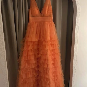 Orange balklänning - Säljer nu min balklänning som enbart testats. Storlek EU38  Pris på hemsida inklusive frakt 1830kr Mitt pris 1000kr  Väldigt tjockt och styvt tyg  Mått: Byst, 90cm  Midja, 72cm  Höft, 98cm Kjol, 150cm  Längd: Golv längd  Tyg: Tyll  Siluett: A-linjeformat  Remmar: Ärmlös  Urringning: V-ringning  Baksida: Dragkedja baktill  Midja Stil: Naturlig  Inbyggd BH: Ja  