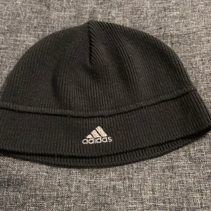Svart stickad mössa från Adidas - Svart ribbstickad mössa från Adidas med broderad logga framtill och diskreta grå detaljer på sidan. Klassisk och enkel design som passar perfekt till kyliga dagar.