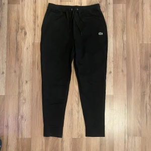 Svarta mjukisbyxor från Lacoste - Svarta mjukisbyxor från Lacoste med klassisk passform och dragsko i midjan. Byxorna har resår vid bensluten och en liten broderad Lacoste-logga på ena benet. Perfekta för en avslappnad stil.