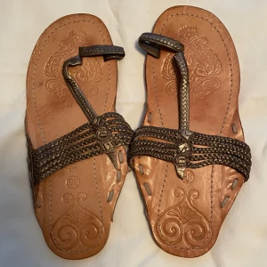 Bruna skinnsandaler med flätade detaljer i stl 38  - Än är inte säsongen över, kanske ska du på solresa i höst/vinter! Använda men i gott skick, se bilder. Säljer ett par bruna sandaler med flätade remmar och dekorativa mönster på sulan. Skorna har öppen tå och platt sula, perfekta för varma dagar. Snyggt hantverk och unika detaljer framtill. Ord pris 299 sek Pris nu 25 sek  