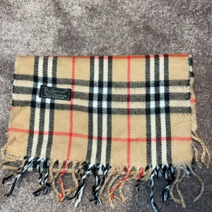  Burberry halsduk - Säljer en klassisk rutig halsduk från Burberry i beige, svart och röd. Den har fransar i ändarna och är perfekt för att hålla dig varm och stilren under kyliga dagar.