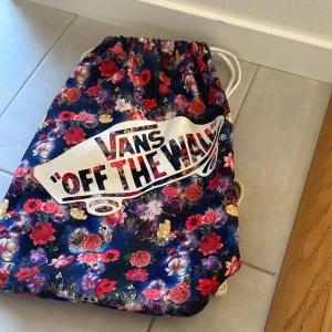Blommig gympa påse från Vans - Säljer en färgglad gympa påse  från Vans med ett blommigt mönster i rött, rosa och blått. Den har också det ikoniska 'Off The Wall'-trycket på framsida 