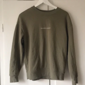 Olivgrön sweatshirt med text - Säljer en olivgrön sweatshirt med texten 'PEACEFUL HEART' på bröstet. Tröjan har en rund halsringning och långa ärmar. Perfekt för en avslappnad stil. Strl S