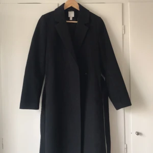 Svart damkappa strl 38 - Elegant svart kappa med långärmad design och bälte i midjan. Perfekt för kyligare dagar med sin klassiska och stilrena look. Köpt från H&M och i strl 38. 