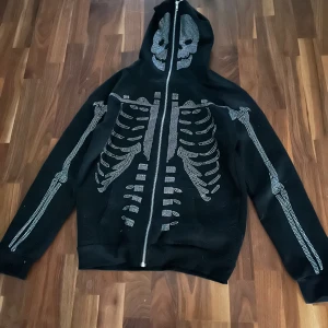 Rhine stone hoodie - Cool svart hoodie med ett detaljerat skelettmönster i vitt som täcker framsidan och ärmarna. Dragkedja framtill och huva med skelettdetaljer. Perfekt för en unik stil.   Ny pris 800kr