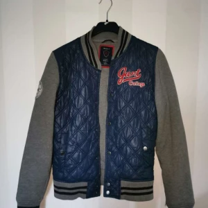 Blå och grå quiltad jacka från Gant - Säljer en blå och grå quiltad jacka från Gant. Jackan har långa ärmar och college-stil med broderad logga på ärmen. Den har knappar framtill och ribbade muddar vid ärmslut och nederkant.