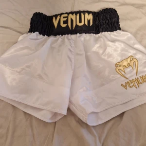 Vita Muay Thai-shorts från Venum - Snygga vita Muay Thai-shorts från Venum med svart och guld detaljer. De har en elastisk midja och Venum-loggan fram. Helt I nyskick, aldrig använt dom.