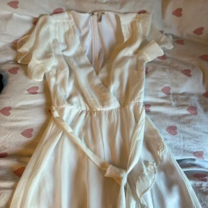 Vit omlottklänning - Fin vit omlottklänning med v-ringning och knytband i midjan. Använd 1 gång på en skolavslutning. Ser gul ut på bilden men det är ljuset, den är vit.