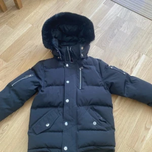 Svart dunjacka från Moose Knuckles - Säljer en svart dunjacka från Moose Knuckles med pälskantad huva. Jackan har en broderad logotyp på insidan och metalliska detaljer på ärmen. Perfekt för kalla dagar! Orginal priset är 14 500kr