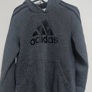 Grå hoodie från Adidas - Mörkgrå hoodie från Adidas med svart logga och klassiska ränder på axlarna. Tröjan har huva, lång ärm och en stor magficka framtill. Perfekt för en sportig och avslappnad stil.