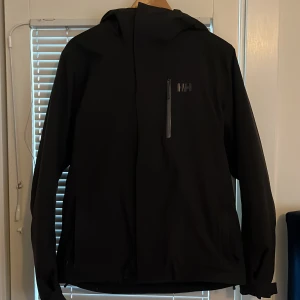 Svart vindjacka från Helly Hansen - Svart vindjacka från Helly Hansen med Luva och diskret HH-logga på bröstet och ryggen. Jackan har dragkedja framtill, bröstfickan med vattentät dragkedja och justerbara ärmslut. Perfekt för blåsiga dagar och stilren look, jackan går även att använda på vintern med en varm kofta under vilket jag har gjort ett par gånger, Storlek S men passar perfekt Med m också. Jackan är köpt av helly Hansen då en ny ligger på 4200. Säljer den för 1200 priset kan diskuteras, är inte omöjlig o förhandla med.