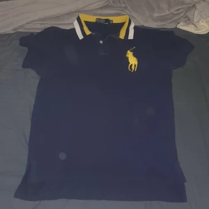 Mörkblå pikétröja Polo Ralph Lauren - Snygg mörkblå pikétröja från Polo Ralph Lauren med stor gul broderad logga på bröstet och gul/vit krage. Klassisk passform och korta ärmar, perfekt till sommaren. Tillverkad i bomull för en skön känsla.
