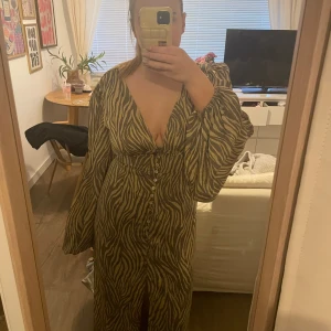 Adoore Klänning - Säljer en långklänning med zebramönster i beige och mörkbrunt. Klänningen har djup v-ringning, långa puffärmar och knappar framtill. Sitter snyggt i midjan och har en lätt slits nedtill. Perfekt för dig som vill sticka ut med ett coolt mönster. Storlek 42, lite liten I storleken, jag brukar ha 38-40!