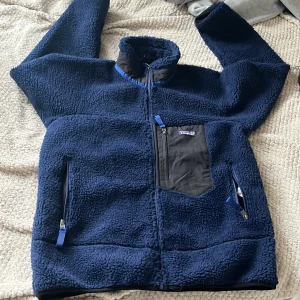 Blå teddyjacka från Patagonia M - Mysig mörkblå teddyjacka från Patagonia i storlek M. Jackan har hög krage, svarta detaljer på bröstfickan och dragkedjor på fickorna. Perfekt för kalla dagar och har en riktigt skön, fluffig känsla. Snygg och funktionell med klassisk Patagonia-logga.