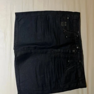 Svarta mini skirt från G-Star RAW - Snygga svarta mini skirt från G-Star RAW, modell 3301. Den har flera coola fickor med knappar och dragkedjor, samt en tydlig vit patch med logga bak. Perfekta för dig som gillar streetwear och vill ha en stilren look.