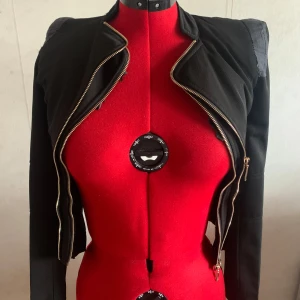 Svart kavajjacka från Qed London - Snygg svart kavajjacka från Geo London med markerade axlar och coola dragkedjor framtill. Jackan är figursydd och har lång ärm, perfekt för dig som vill ha en edgy och stilren look. Materialet känns stabilt och har en lätt stretch. Har lite slits på insidan men inget som påverkar användningen 