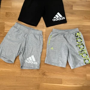 Adidas shorts i svart och grått - Tre par Adidas shorts i sportig stil. Ett par är svarta med stor vit Adidas-logga, de andra två är gråa – ett med vit logga och ett med gulmönstrad Adidas-tryck och gul detalj vid fickan. Alla har resår i midjan och är perfekta för träning eller chill. Svarta adidas shortsen och grå är det 164 och den tredje adidas shortsen med grön logga är 154. 30kr styck men för alla tre 80kr
