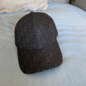 Dior keps - Snygg svart keps från Dior med klassiskt monogrammönster över hela kepsen. Justerbar baktill och fodrad med Dior-logga på insidan. Perfekt accessoar för att lyfta din outfit med en lyxig touch.