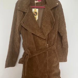 Brun manchesterjacka från Bubbly Moda - Snygg brun manchesterjacka från Bubbly Moda med bred krage, långa ärmar och bälte i midjan. Jackan har markerade vertikala ränder och dragkedja framtill. Perfekt för dig som vill ha en trendig och bekväm jacka med cool retrovibe.