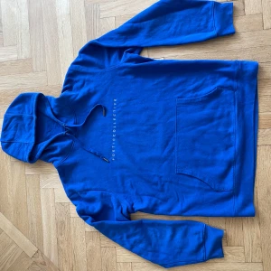 Blå hoodie Poetic Collective XL - Blå hoodie från Poetic Collective i storlek XL. Framsidan har vit textlogga och baksidan har en stor färgstark grafisk print i blått, rött, vitt och svart. Tröjan har huva med snörning och en klassisk magficka. Perfekt för dig som gillar streetwear och konstnärlig stil.